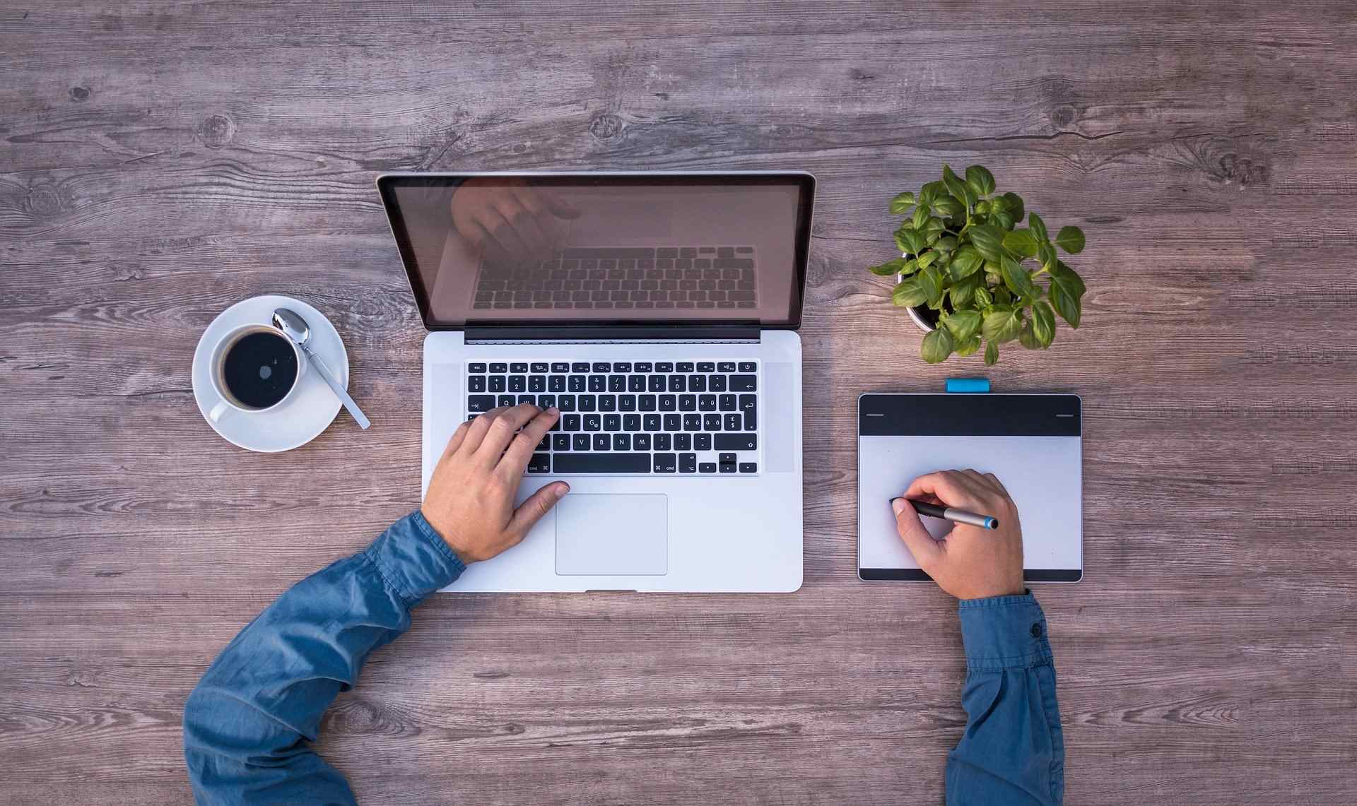 POV auf einen Schreibtisch mit Laptop, Kaffeetasse und Mitarbeiter